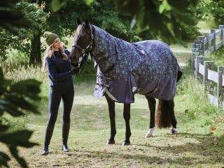 A horse wearing a floral print rug stands in a field with a girl.