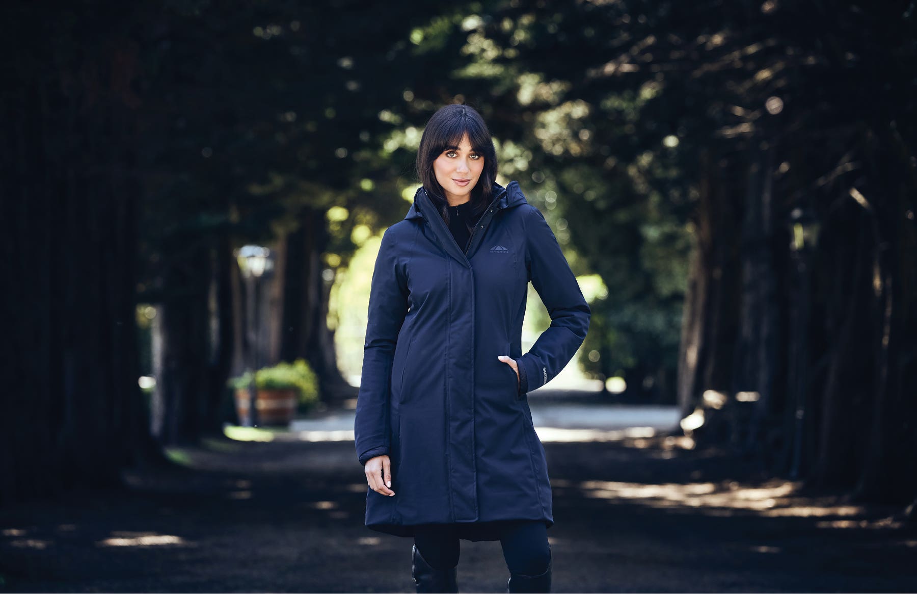 Model standing in tree lined laneway wearing WeatherBeeta All Purpose Longline Waterproof Equestrian Jacket